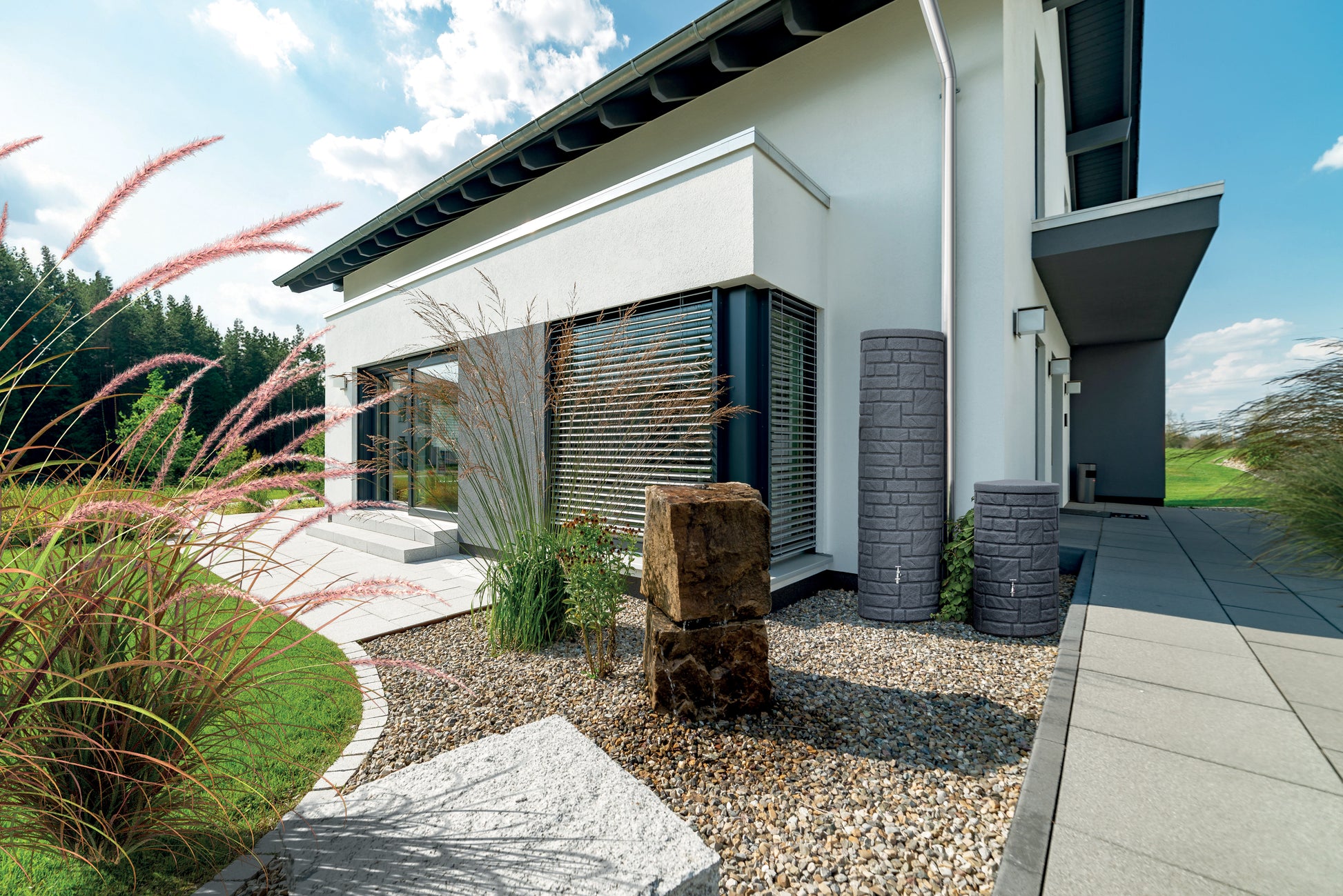 Moderne Hausfassade mit angelegtem Garten, Zierkies und einer YourCasa Regentonne [StoneRain] – frostsicher & UV-beständig.