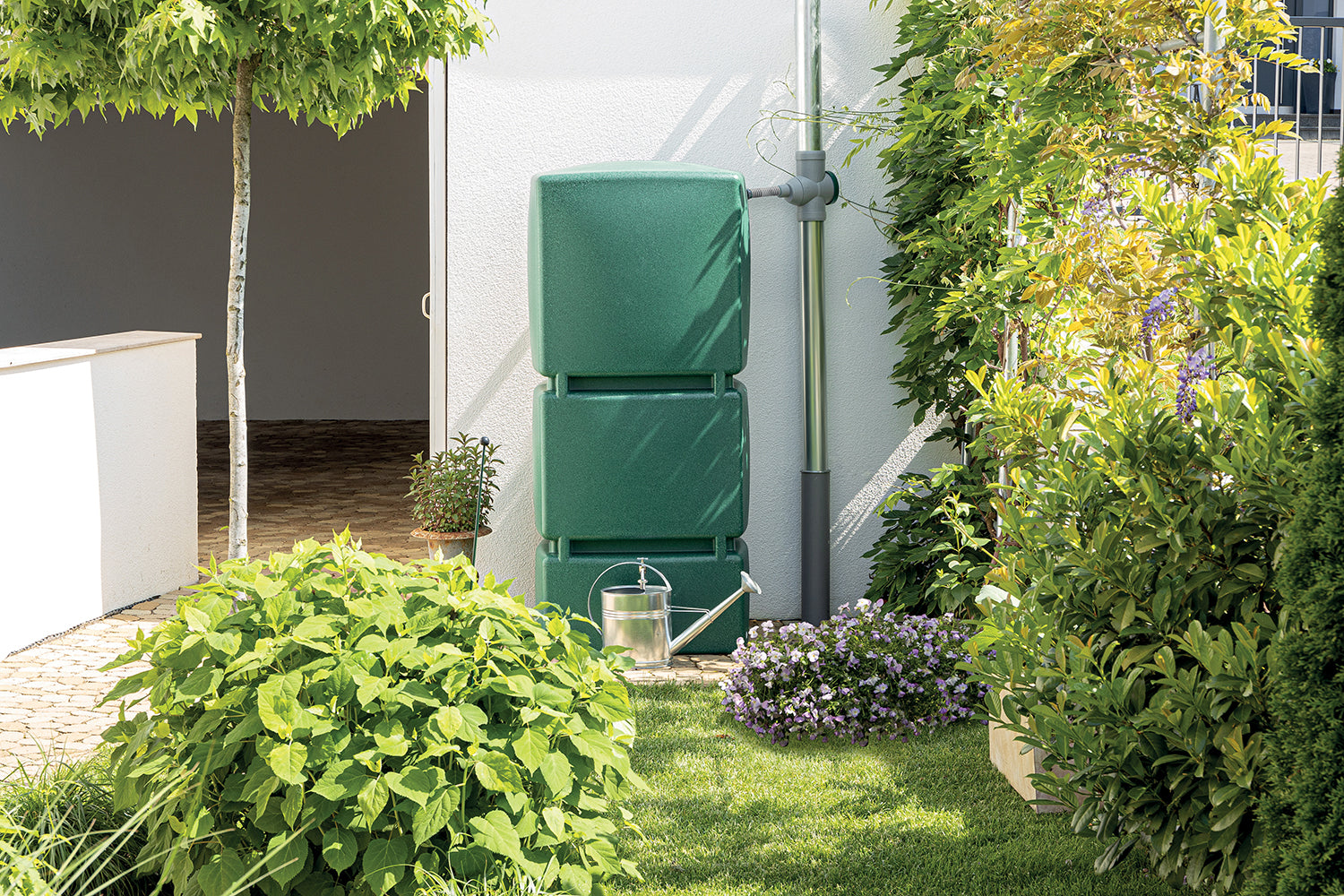 YourCasa® Regensammler [Downpipe70] - Mit Überlaufschutz + Anschlussschlauch 32mm Regentonne an einem Haus neben blühenden Pflanzen und einer Gießkanne installiert.