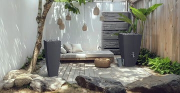 Eine minimalistische Terrasse besticht durch ein weißes Sofa, ein Holzdeck, einen Steinweg und graue Pflanzkübel, in die sich eine YourCasa Regentonne von yourcasa-de nahtlos einfügt. Sie ist frostbeständig und UV-stabil und ergänzt Hängelampen und üppiges Grün, um die ruhige Atmosphäre zu unterstreichen.