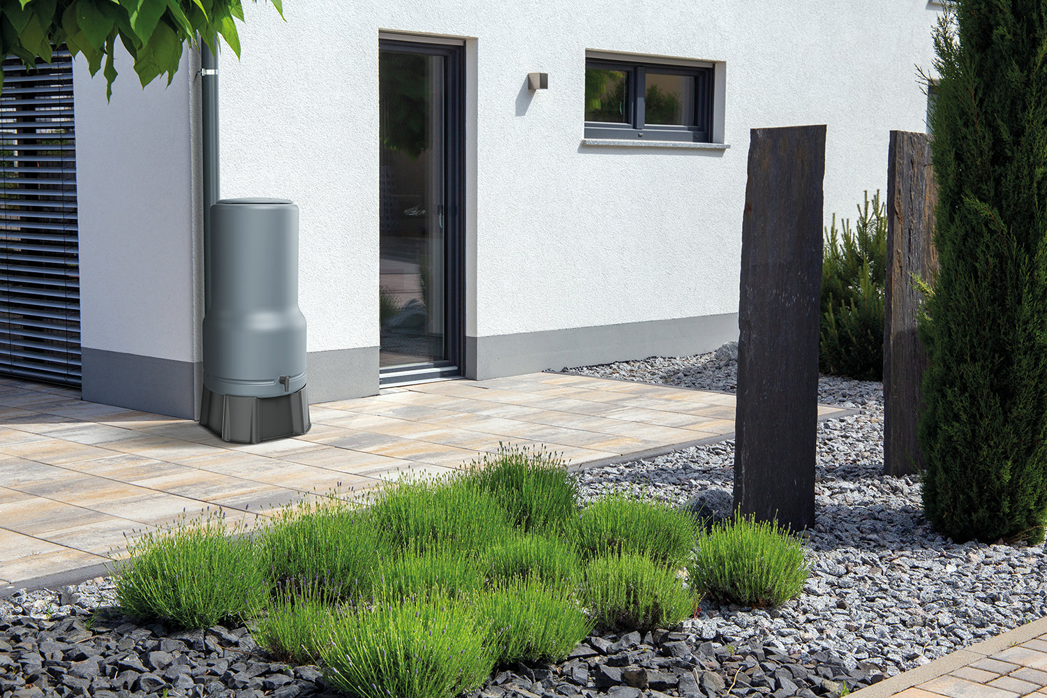 Moderne Hausaußenansicht mit einem angelegten Garten, der eine YourCasa Regentonne [RainBell] mit Standfuß, Deckel und Wasserhahn für nachhaltige Bewässerung, Ziergräser, Kies und Geometrie. Markenname: yourcasa-de