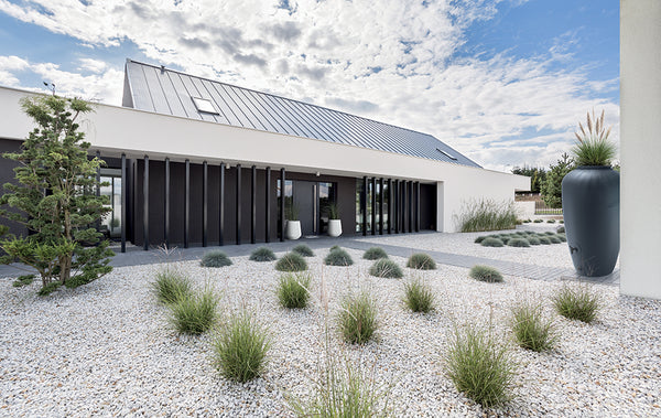 Modernes einstöckiges Haus mit einem angelegten Garten mit Ziergräsern, Kies und einer Yourcasa-de Regentonne 210/360/440 Liter – Amphore Design zur Nutzung von Regenwasser.