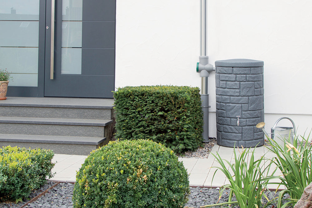 Ein YourCasa Regentonne [StoneRain] mit Metallgewinde und Deckel in Steinoptik - Regenfass Frostsicher aus Kunststoff Grauwassertank vor einem Haus.