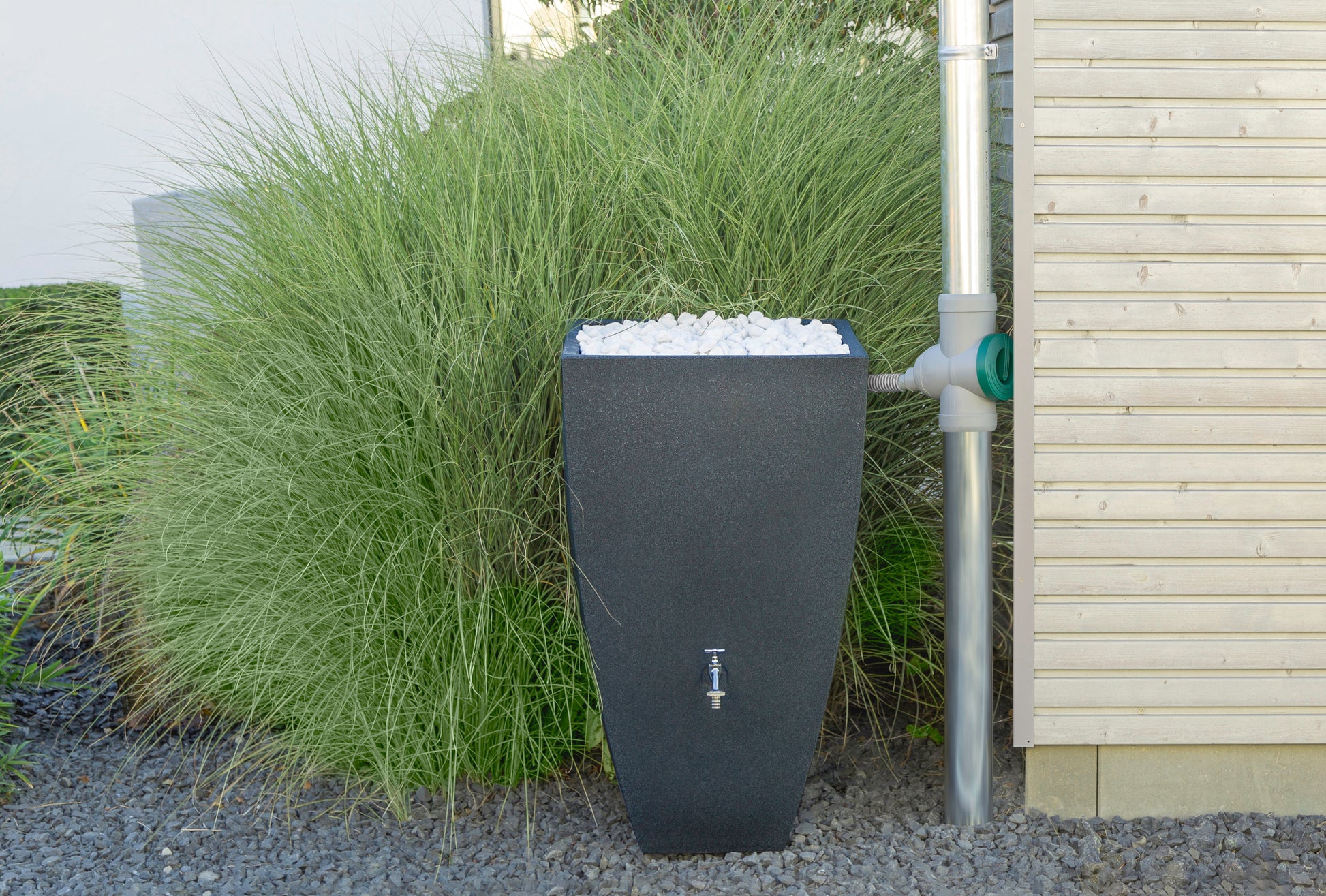 Moderne Regenwasser-Sammeltonne neben Ziergras an einem Haus aufgestellt mit angeschlossenem YourCasa® Anschlussschlauch 32mm für Regensammler inklusive Handbohrer von yourcasa-de.