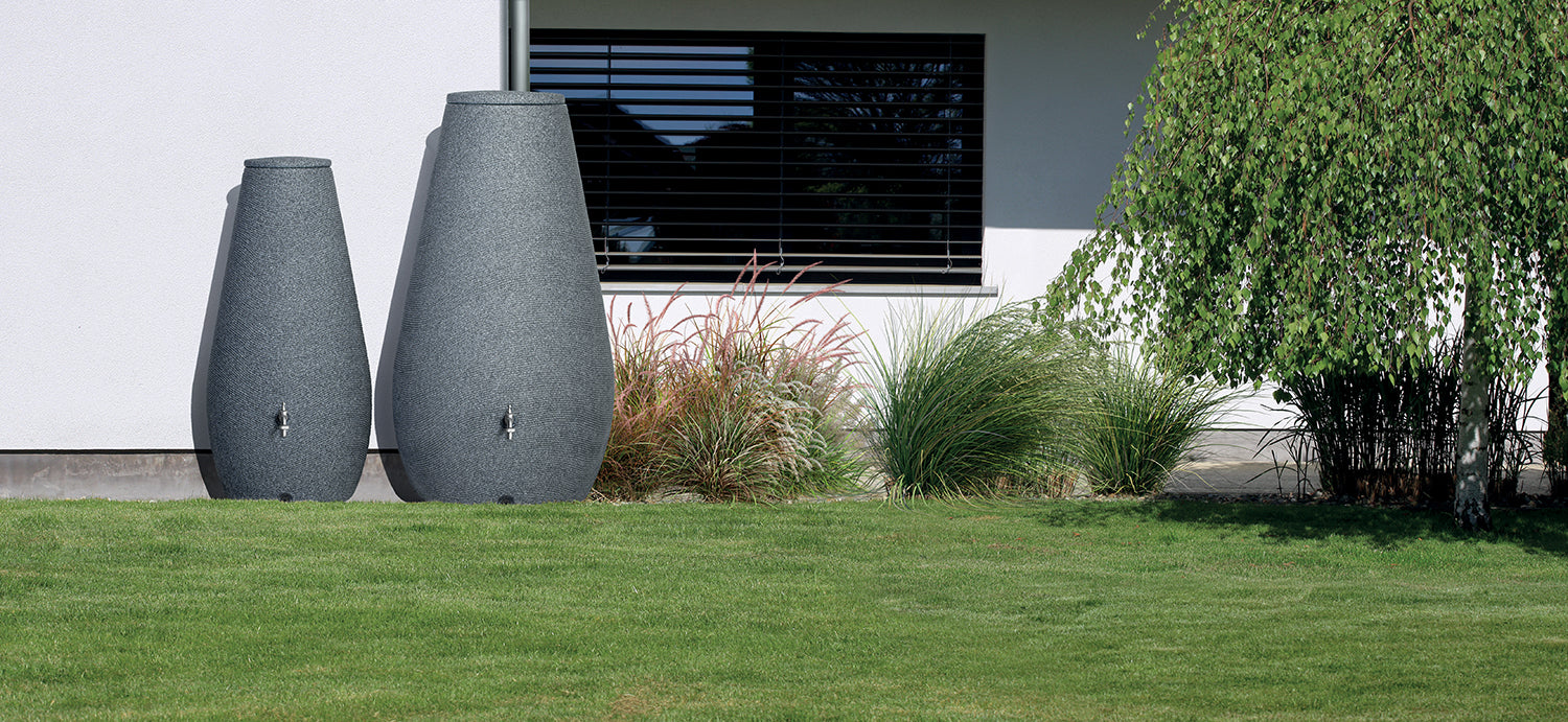 Zwei große Dekovasen neben einem modernen Gebäude mit Ziergräsern und einem Baum erleichtern die Gestaltung von YourCasa Regentonne 200 Liter [EcoDrop] – Nachhaltige Gartenbewässerung von yourcasa-de.
